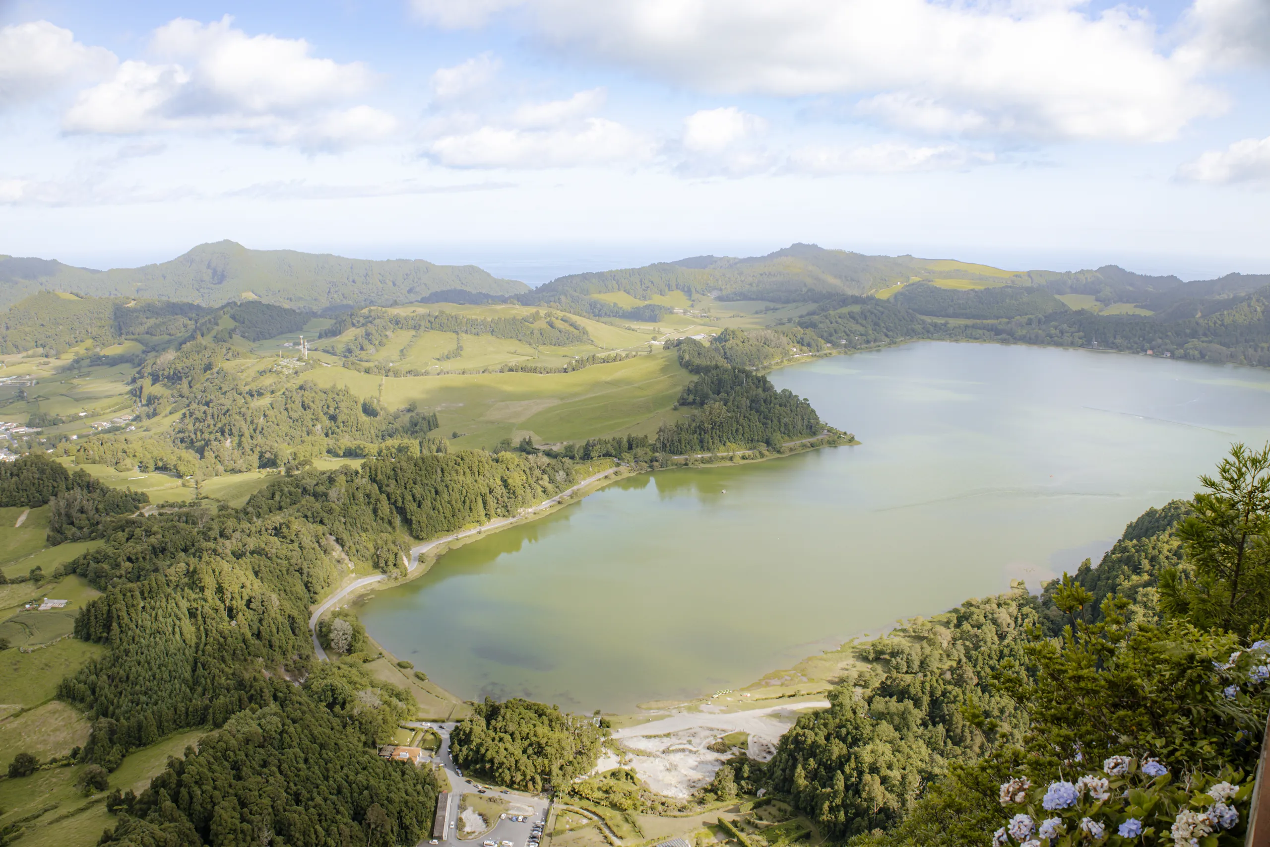 La Lagoa das Furnas vista dall'alto, circondata da boschi e pascoli, con le ortensie azzurre in primo piano e le colline sullo sfondo.