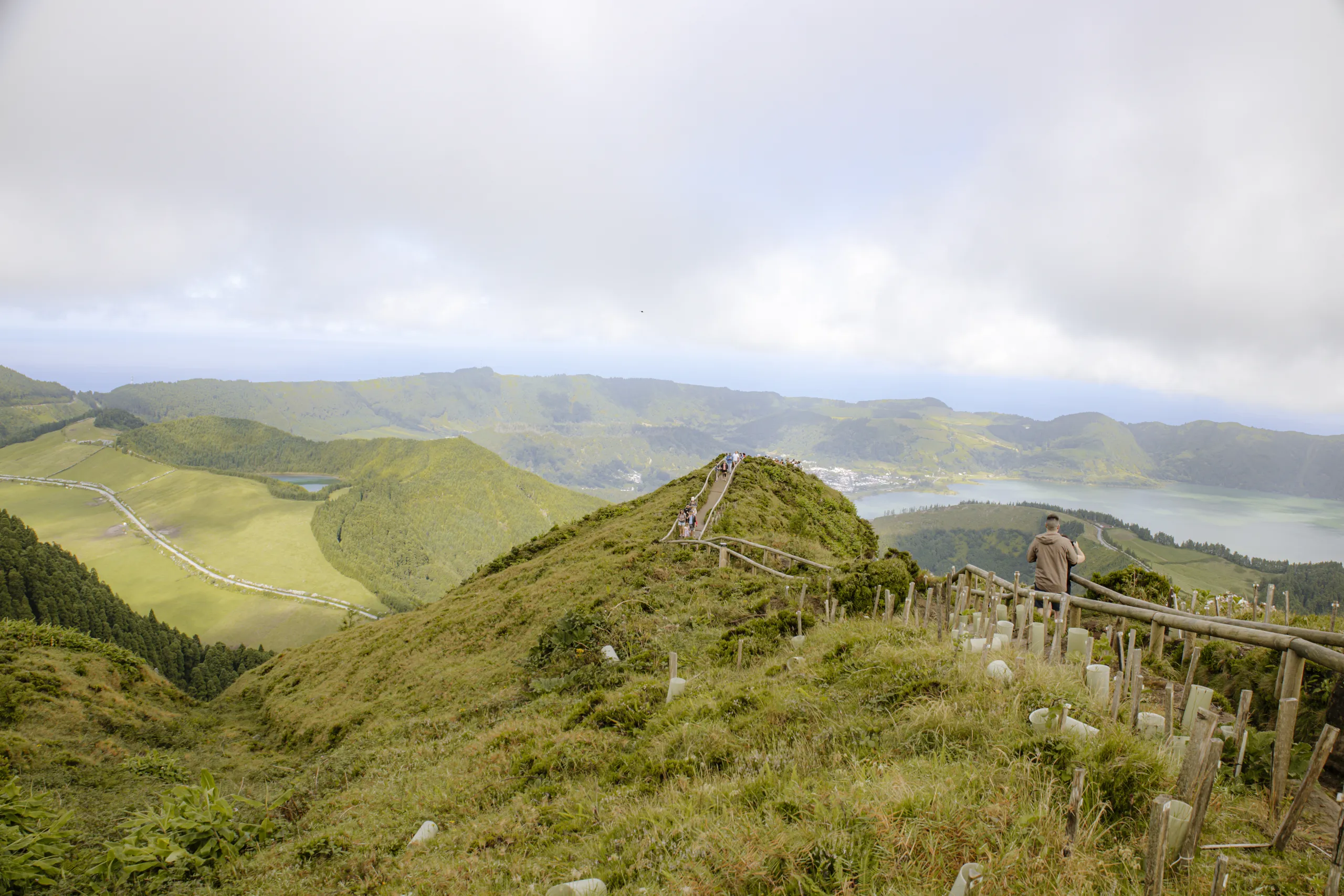 Il sentiero con la staccionata di legno che sale lungo un crinale erboso verso la Grota do Inferno, con i laghi di Sete Cidades e le colline sullo sfondo.
